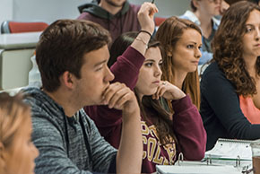 students in a classroom. A girl wearing a Boston College sweatshirt raises her hand in the center of the fram
