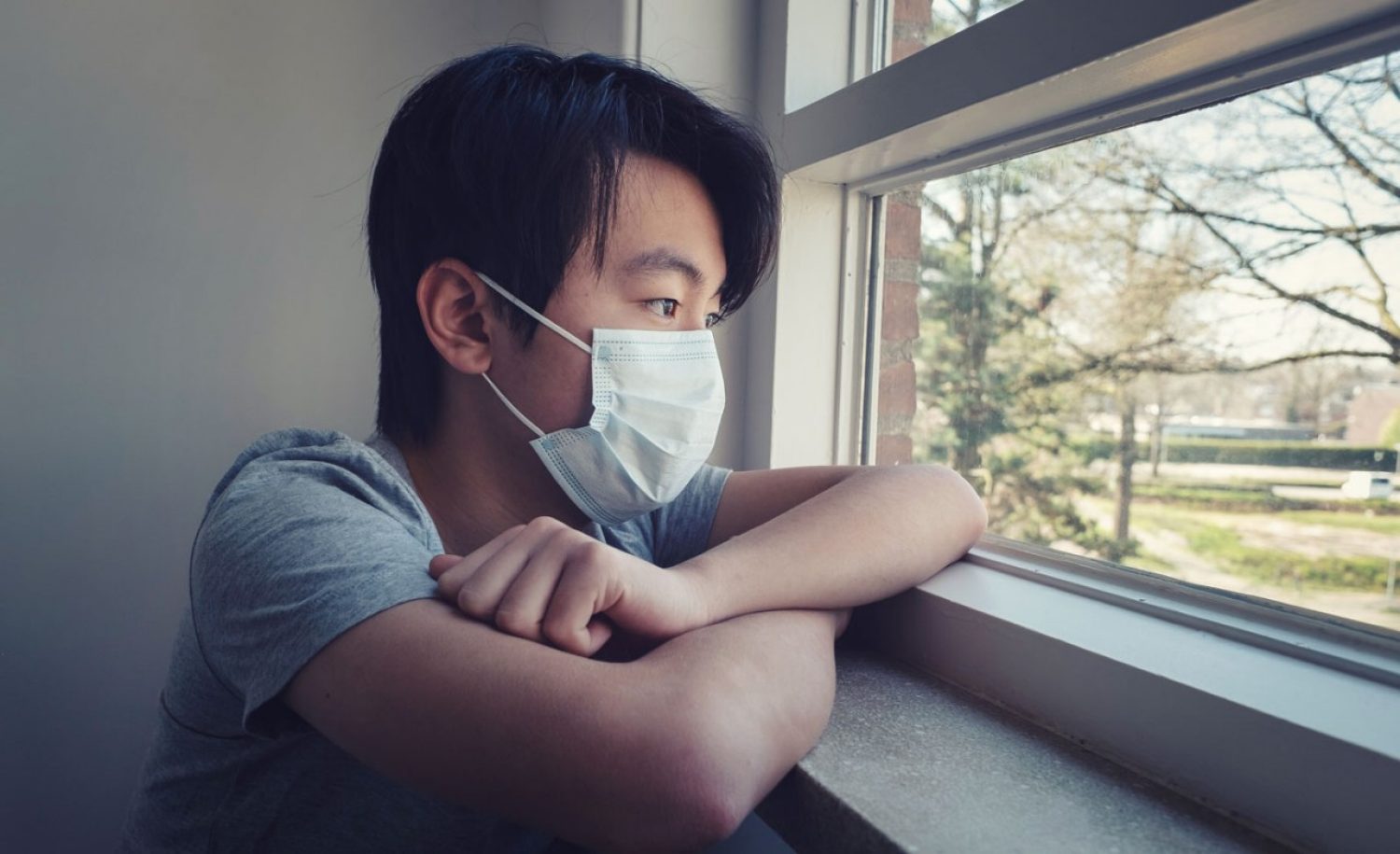 Young man in a medical mask at a window with grass and trees outside