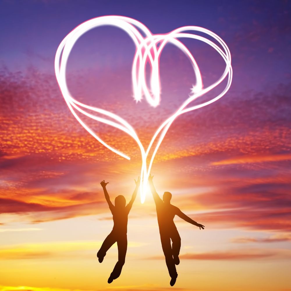 Two people jumping with their arms reached up and the outline of a heart lit between them in the sky