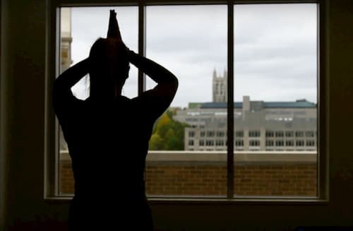 Silhouette of person with prayer hands at forehead, looking out window towards Gasson Hall