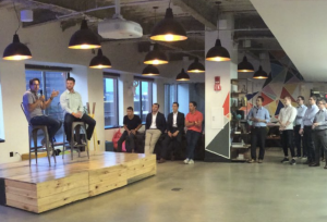 two men sit on stools and speak to a group