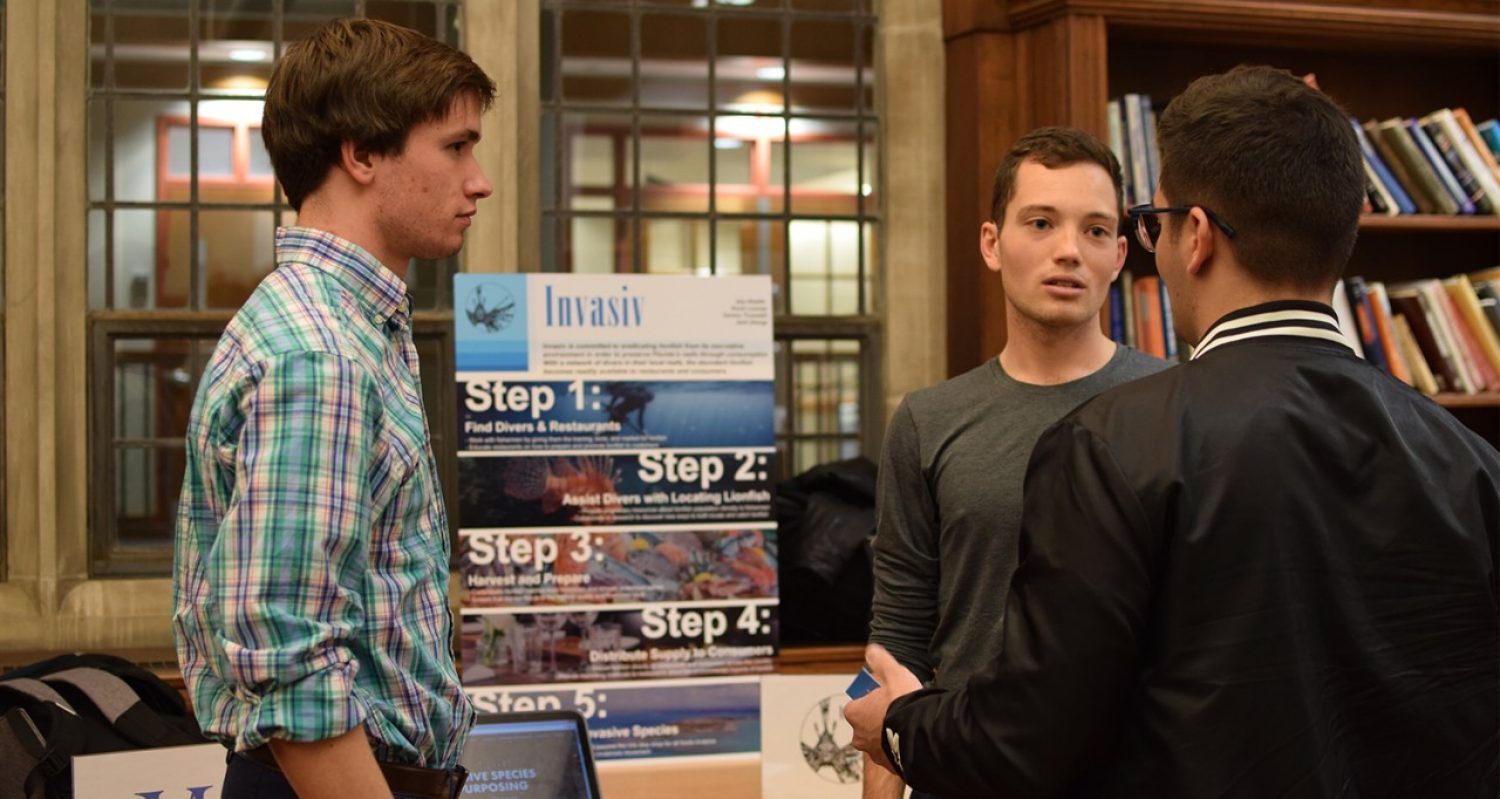 Two students explain their pitch idea to another student front of a posterboard in the Fulton Honors Library