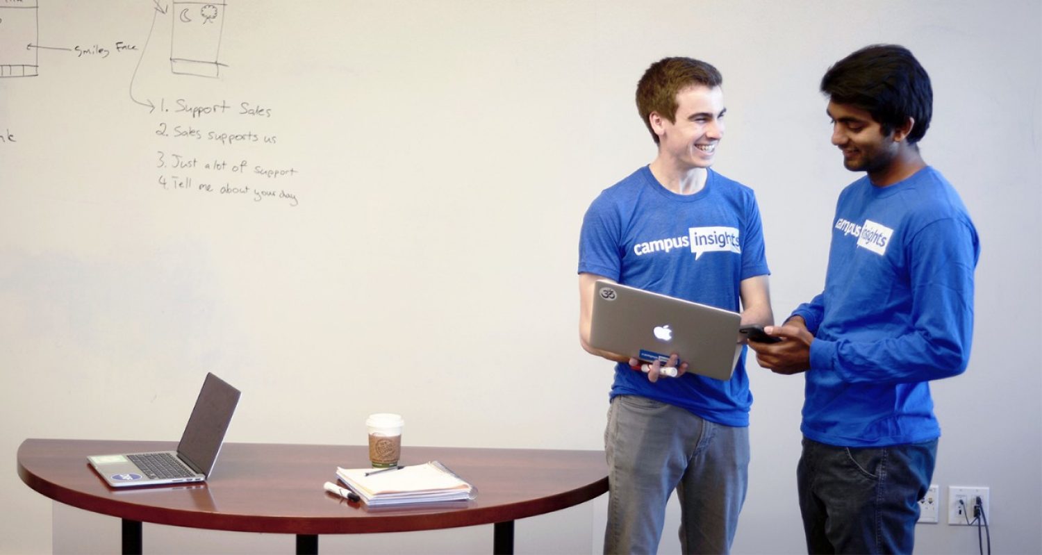 Two young men in blue CampusInsights shirts stand in front of a whiteboard and look at a laptop