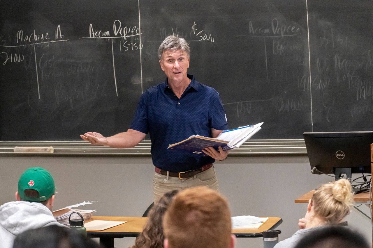 Male professor in a blue collared shirt stands in front of a blackboard with economics terms written on it. His right arm is extended and he holds a book in his left hand. Students sit in front of him