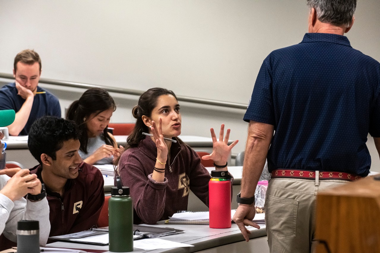 Students sit in a classroom. At the center of the image a student in a BC sweatshirt is talking with her hands gesturing in front of her face. The professor from the previous photo is standing facing her with his back to the camera