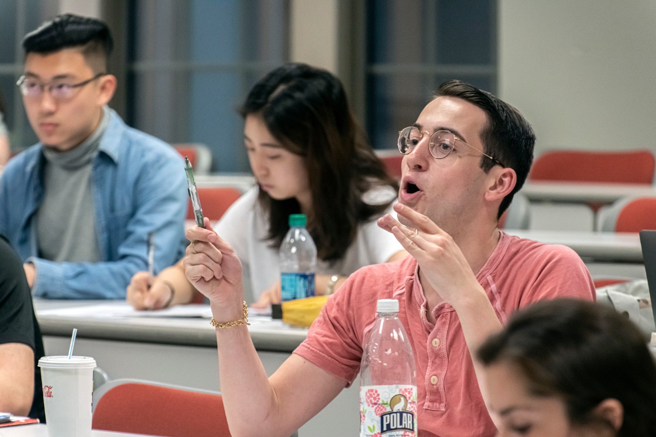 Students sit in a classroom. In the foreground of the image a student with glasses in a salmon colored shirt if gesticulating while holding a pen. His mouth is open as he is speaking