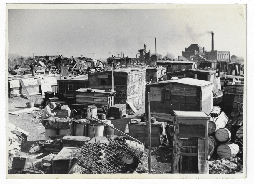 Black and white photo of a dump, which includes a number of dilapidated shacks and barrels.