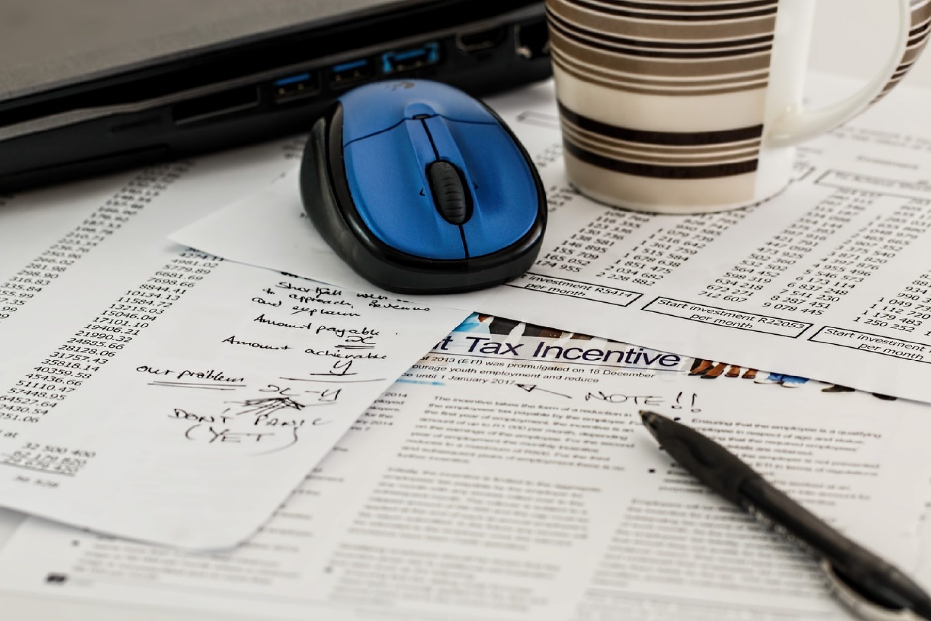 Accountant notes on financial papers with financial numbers partly covered by a pen, coffee mug, blue wireless mouse, and laptop