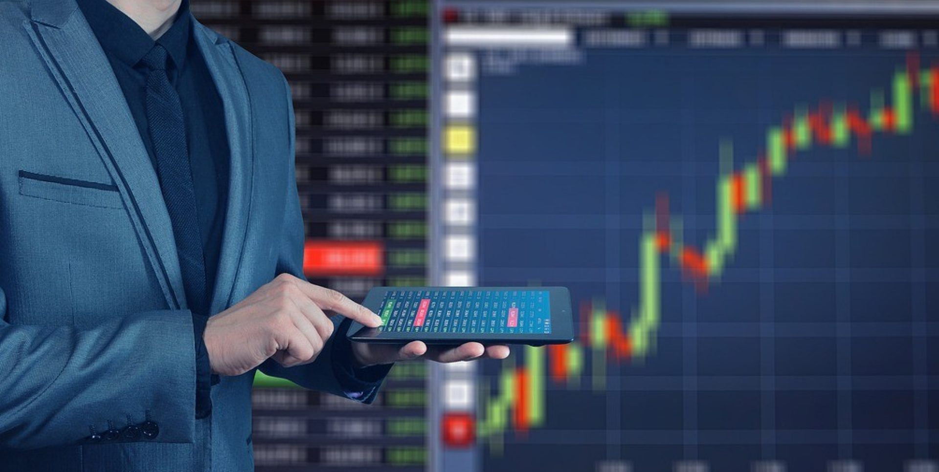 Person wearing a suit standing in front of a screen featuring stock readouts