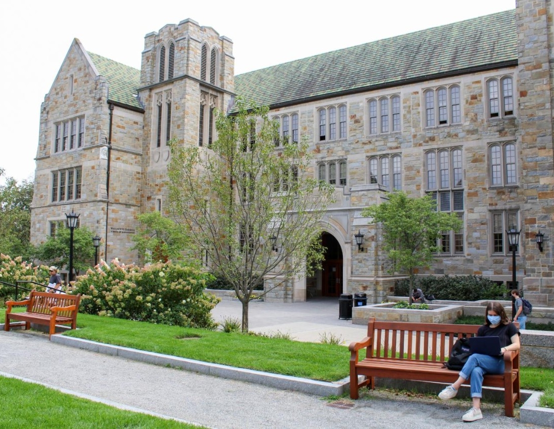 A masked student sits on a bench outside of Fulton Hall, working on a laptop