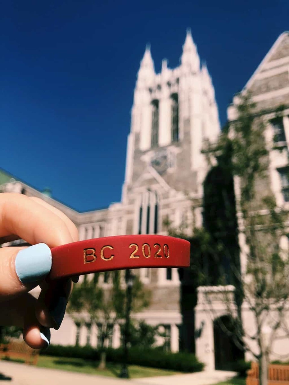 a maroon BC 2020 bracelet is held in front of Gasson Hall