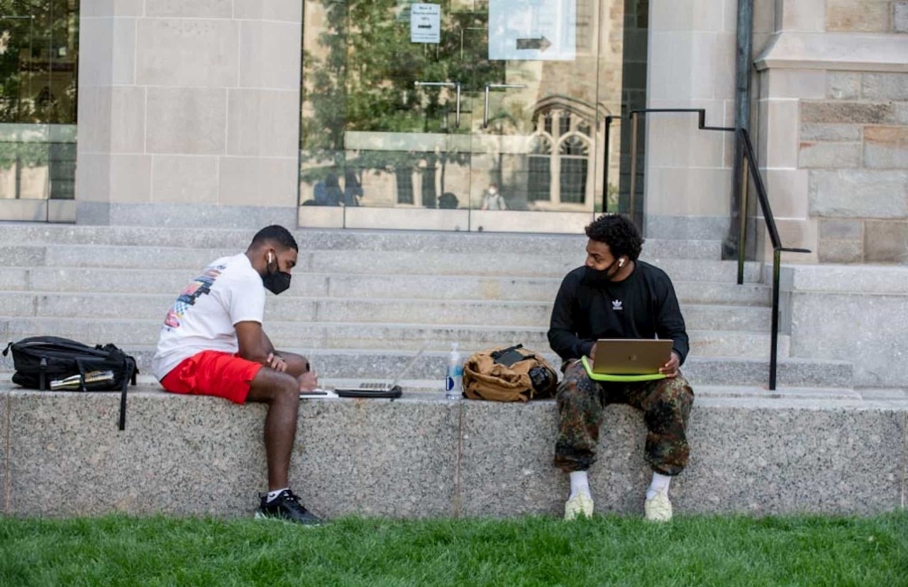 two masked students sit outside with laptops