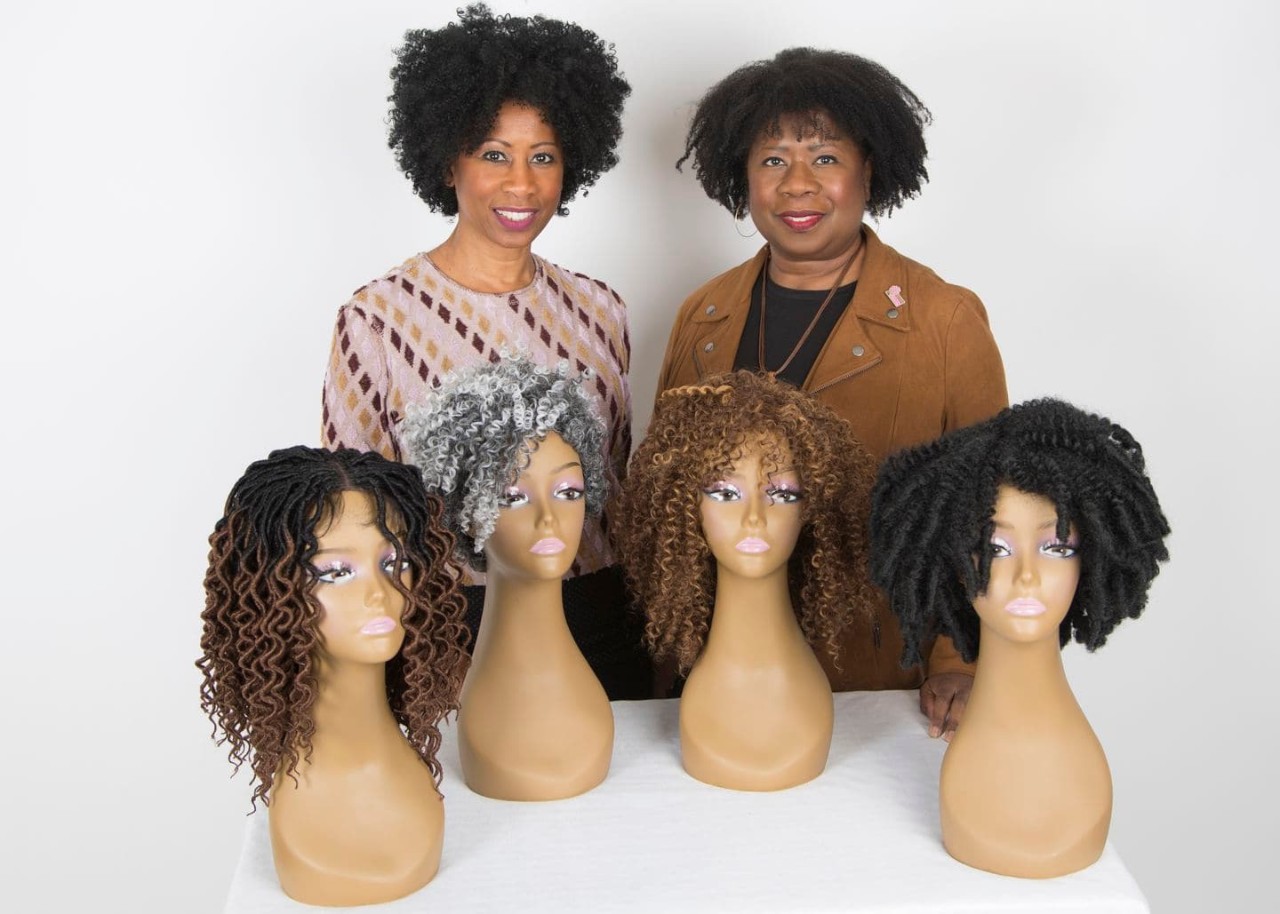 two black women stand behind a table of wigs with textured hair