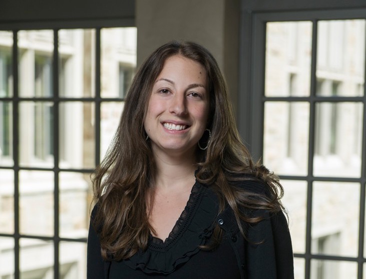 Photo of A woman with long dark hair and wearing a black shirt, smiling and posed in front of windows.