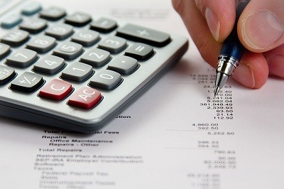 Photo of Close-up image of a calculator and a hand holding a pen, making notes on a piece of paper with numbers on it