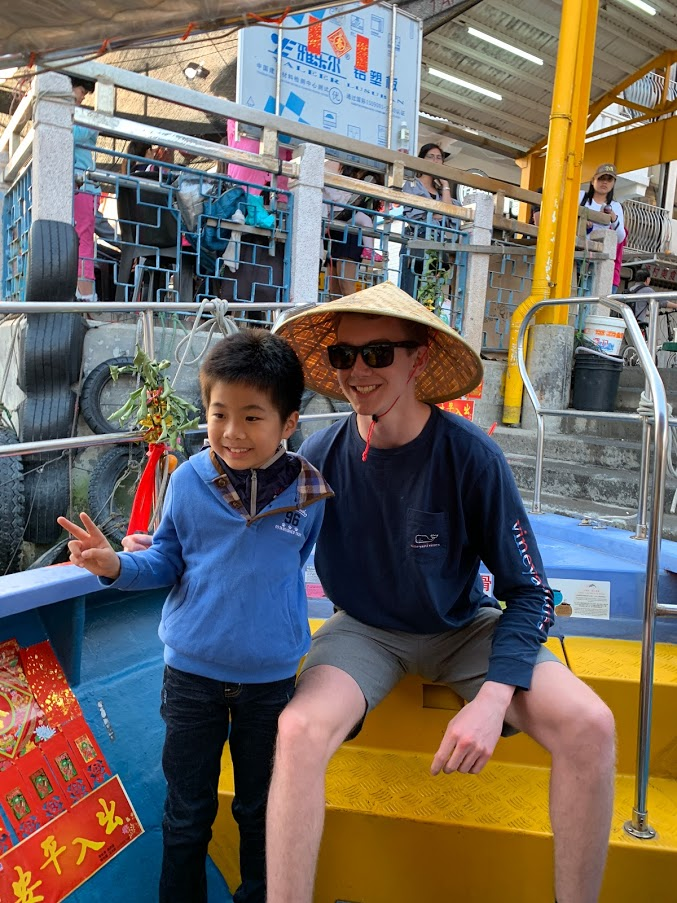 Austin and a young boy in a crowded market. Austin is wearing a traditional Asian rice hat
