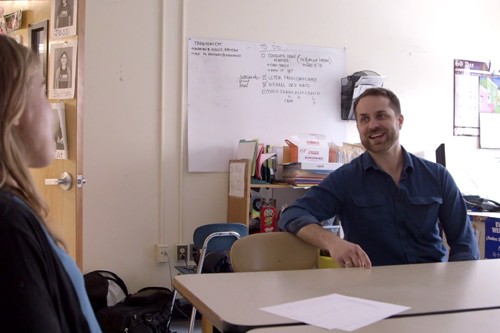 Man in blue dress shirt talks to a woman, both sitting at a table