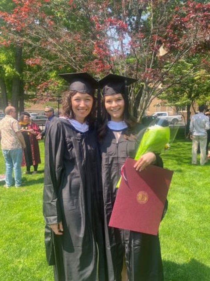 Photo of 2 women stand outside in graduation robes holding diplomas