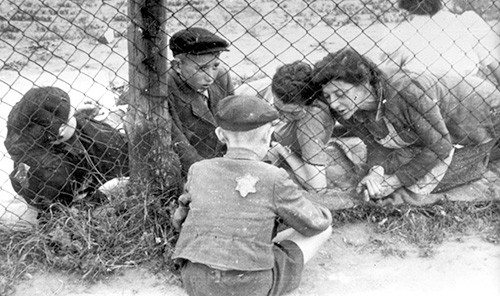 Black and white photo of a woman and children sitting on either side of a chainlink fence, one boy wearing a star of David on his back