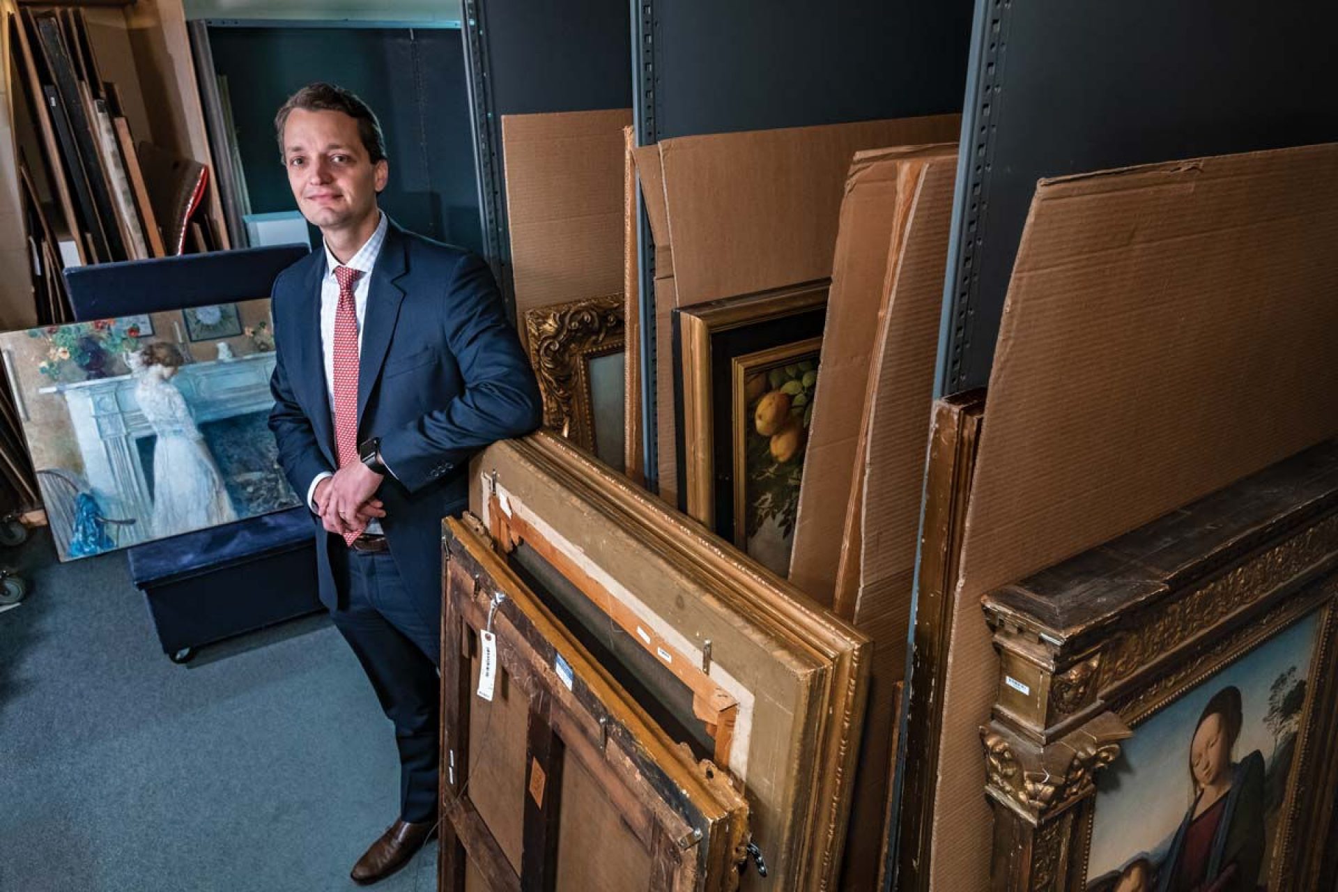 Portrait of Will Haydock in a storage room with paintings around him.