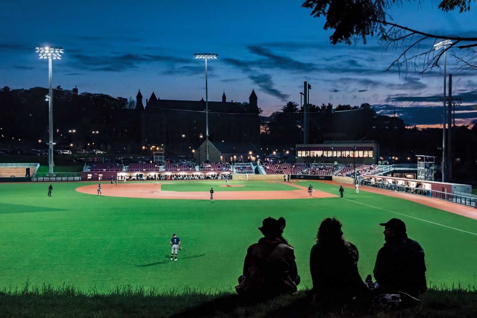 Three fans in silhoette watch a lit up baseball game at dusk