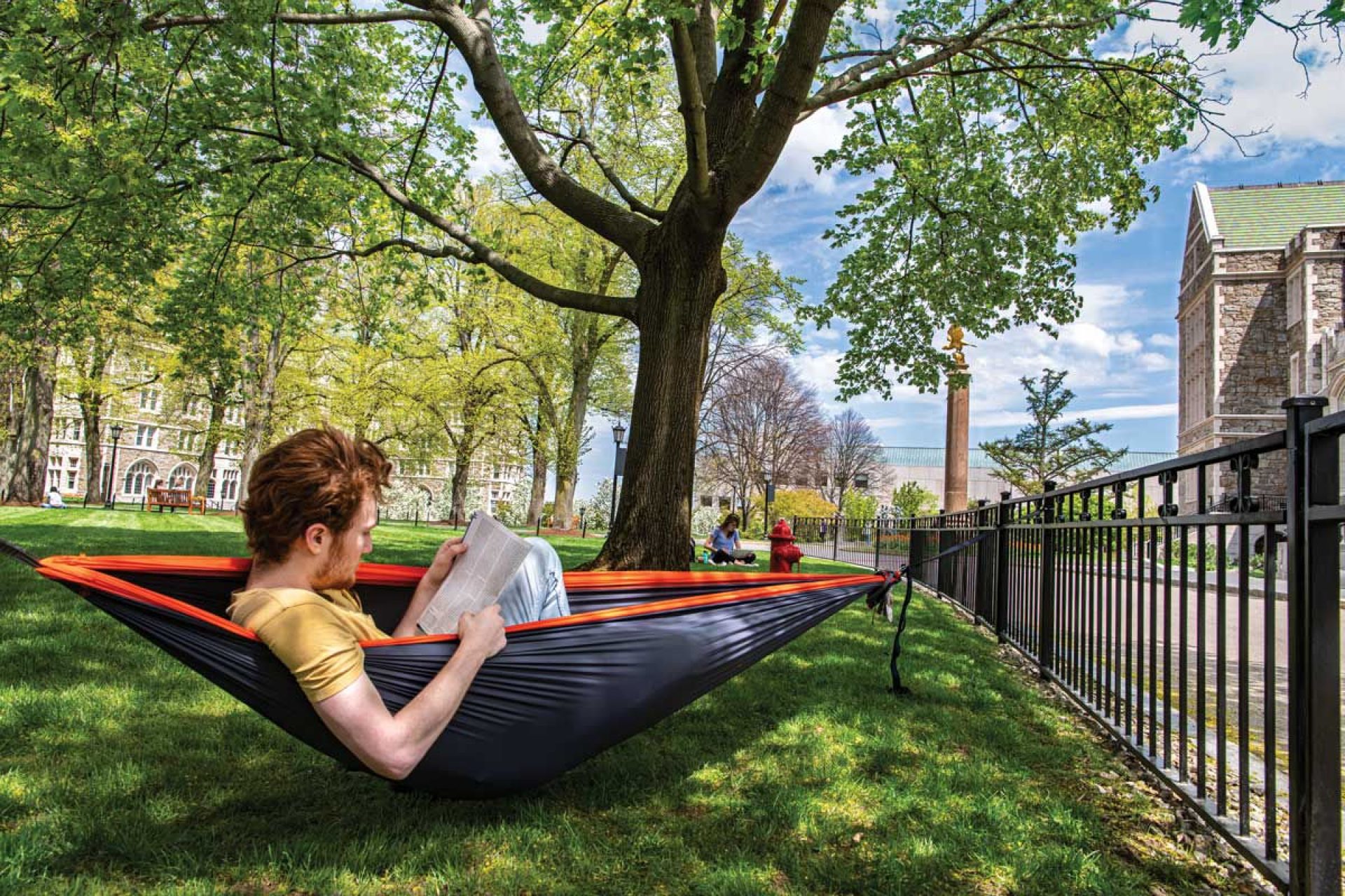 Student reading in a hammock under the trees on a sunny day with Gasson Hall in the background