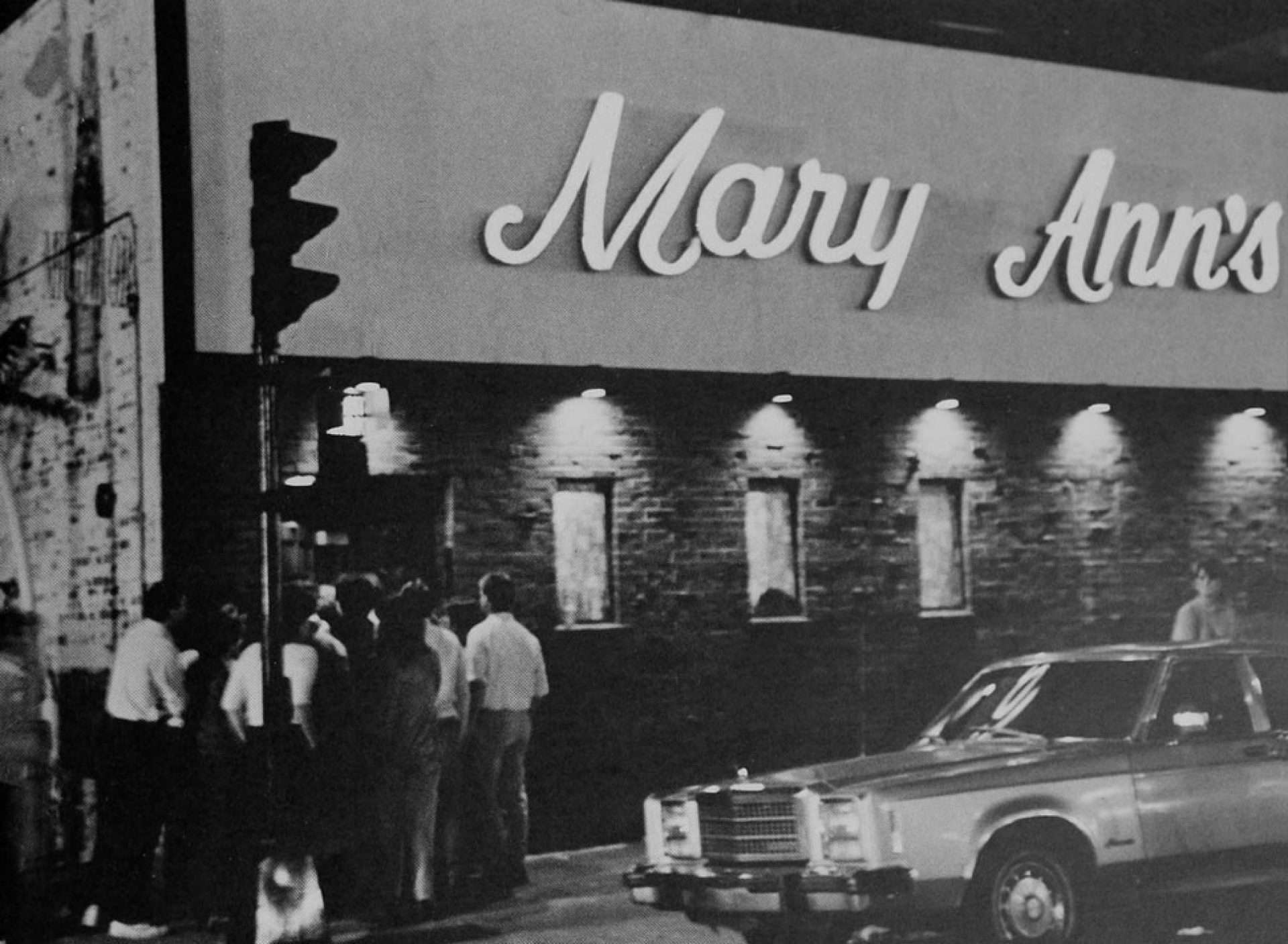 Black and white photograph of people gathered outside a bar in the evening