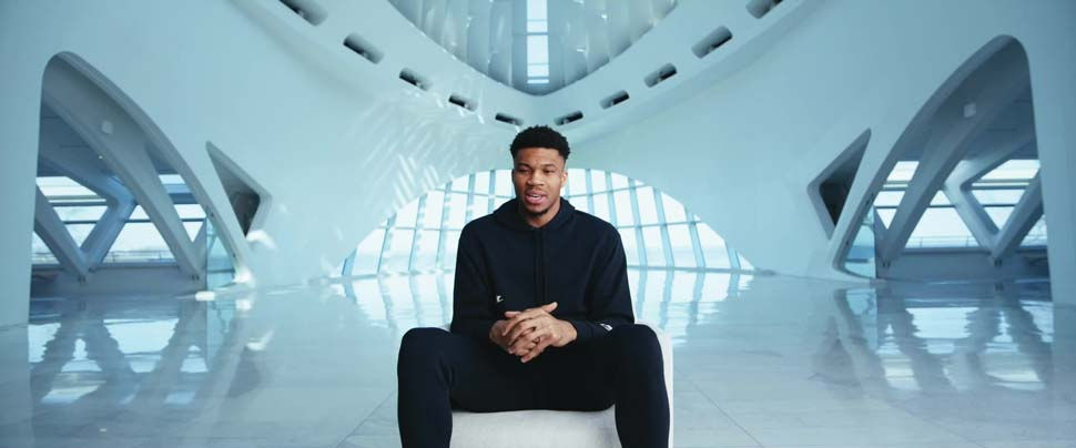 Giannis speaking while seated alone in a massive arched interior.