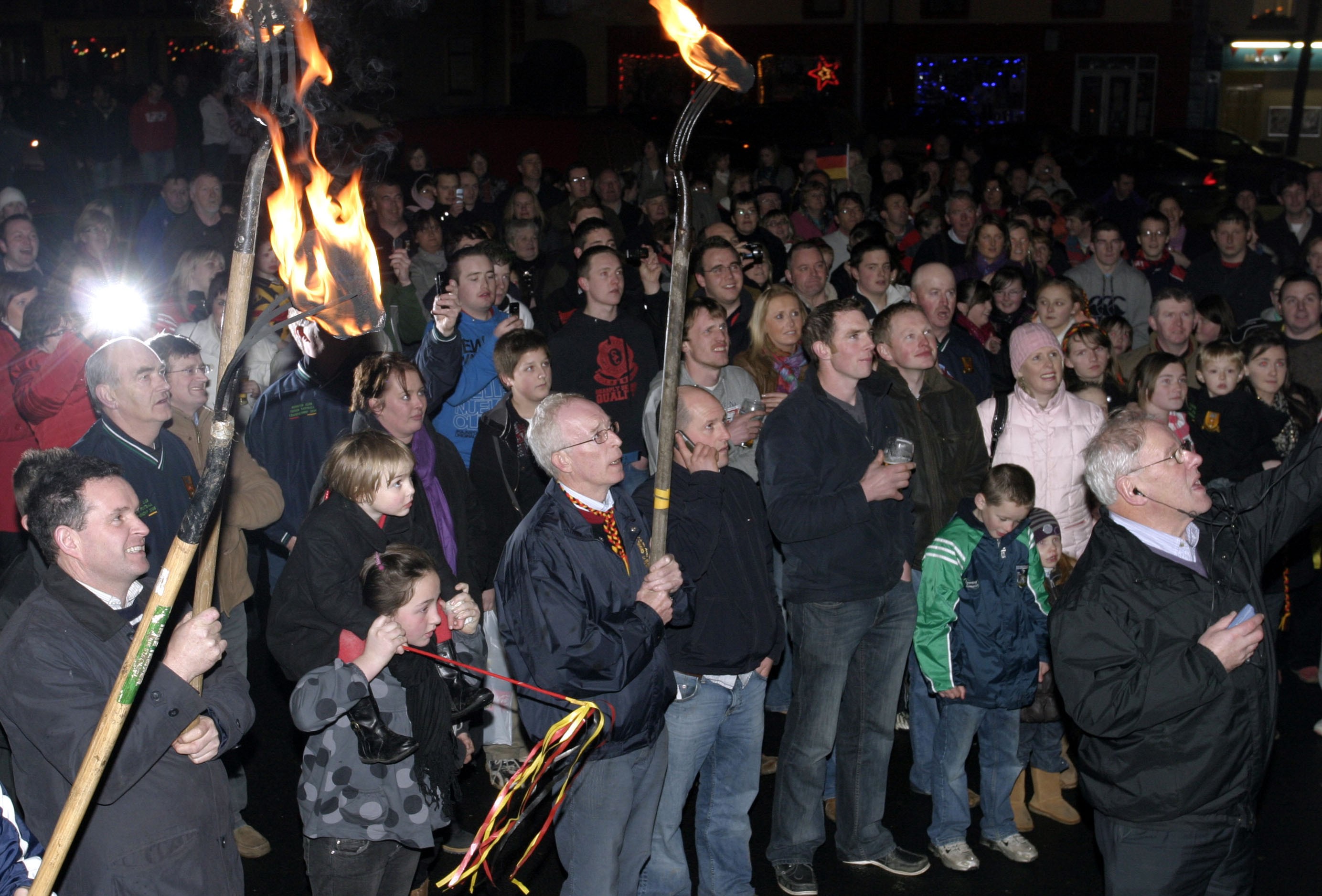 A scene from a Drumcollogher-Broadford GAA victory homecoming celebration. Note the lit turf burning on pitchforks in the foreground. The carrying of lit turf and the burning of bonfires are traditional GAA homecoming celebration rituals.