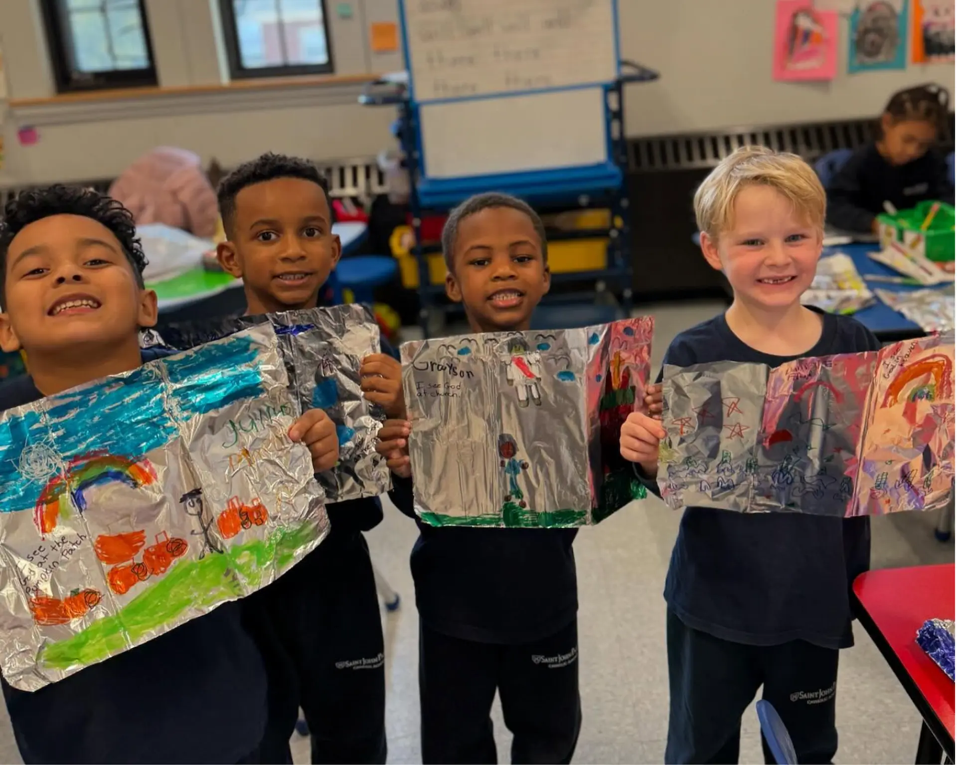 Four smiling young boys hold up drawings of God on tin foil.