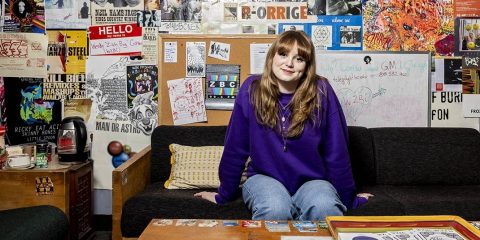 Lily Telegdy sitting on a couch against a wall of posters and stickers