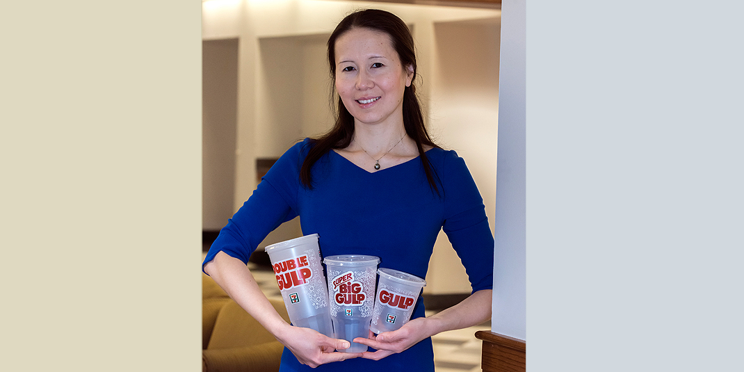 Nailya Ordabayeva in a blue dress, posing with three sizes of fast food soda cups