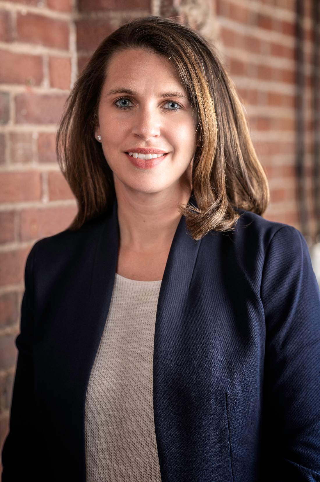 A headshot of Cory Munchbach. She is a woman with a light skin tone and shoulder length brown hair. She has bright blue eyes and she is wearing a blue blazer and a gray shirt. She is smiling at the camera while she stands in front of a brick wall.