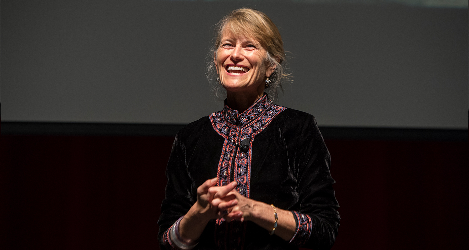 Jacqueline Novogratz standing on a stage, smiling with her hands clasped during a presentation