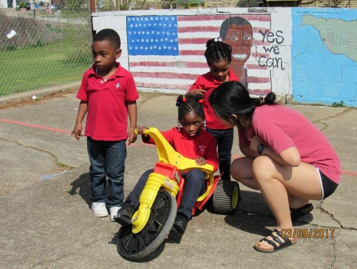 a student crouches next to a young child on a tricycle. two other young children in school uniforms stand nearby