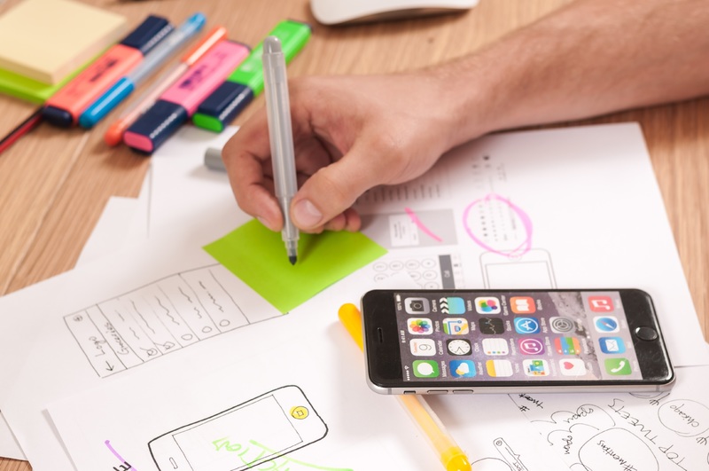 a hand writes notes on a post-it with an iphone in the foreground and diagrams and charts also on the table