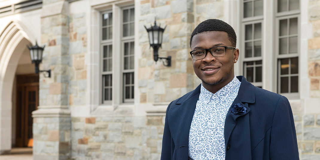 Kofi Nimo, a young black man in glasses and a button down with a jacket, stands in front of Fulton Hall