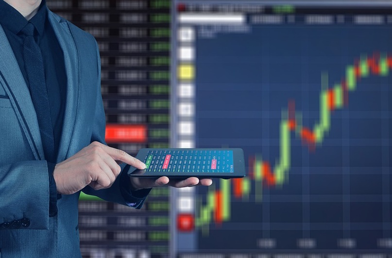 Person wearing a suit standing in front of a screen featuring stock readouts