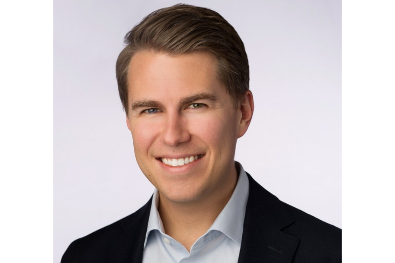 Headshot of Miles Taylor, polished younger man smiling in a button-up shirt and coat.