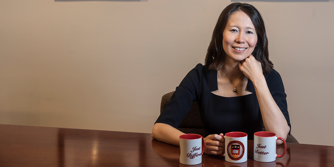 Nailya Ordabayeva sitting at a table. Three mugs are in front of her, two saying "Just Better," and "Just Different," respectively, and one with the Carroll School logo