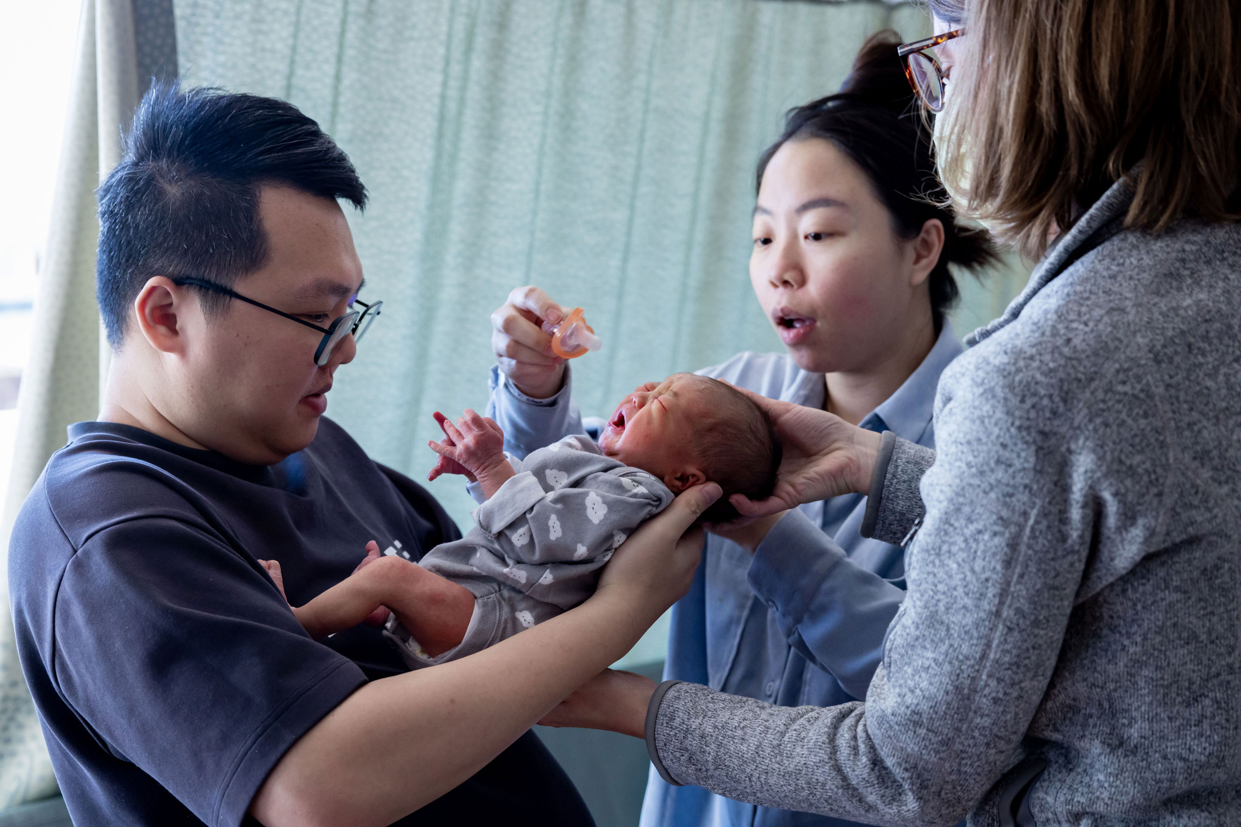 A father holds his crying baby and the mother is placing a pacifier in the baby's mouth. The midwife supports the baby's back and head.