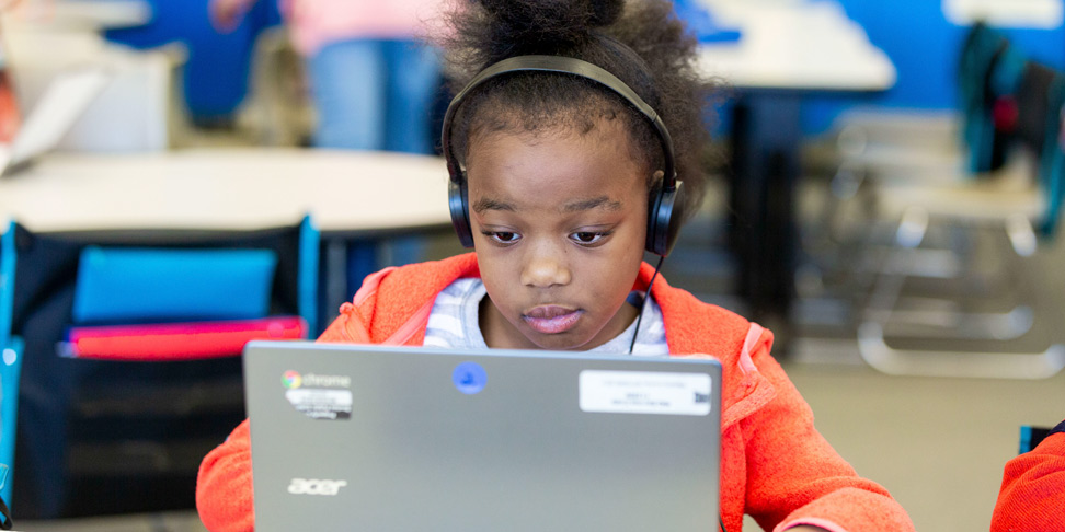 Elementary aged girl in orange sweatshirt with headphones on her ears in front of computer