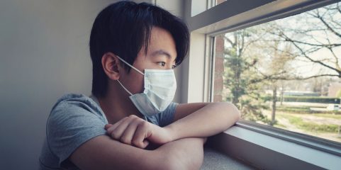 Young man in a medical mask at a window with grass and trees outside