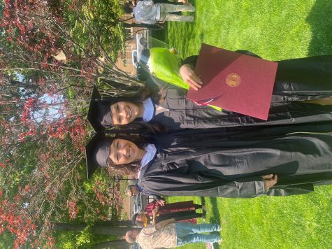 2 women stand outside in graduation robes holding diplomas