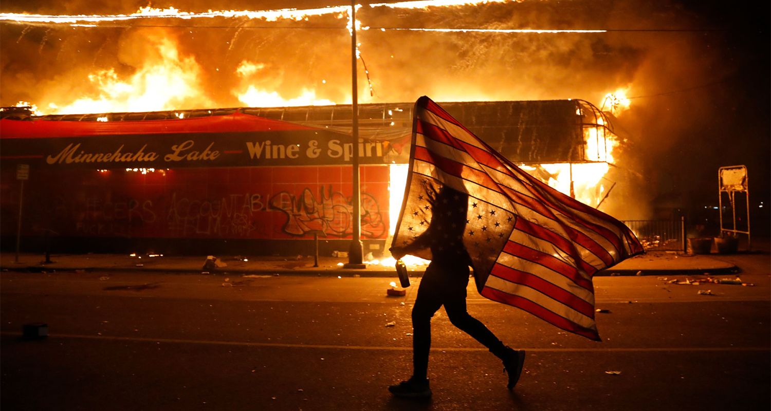 A protester carries a U.S. flag upside, a sign of distress, next to a burning building Thursday, May 28, 2020, in Minneapolis. Protests over the death of George Floyd, a black man who died in police custody Monday, broke out in Minneapolis for a third straight night. (AP Photo/Julio Cortez)