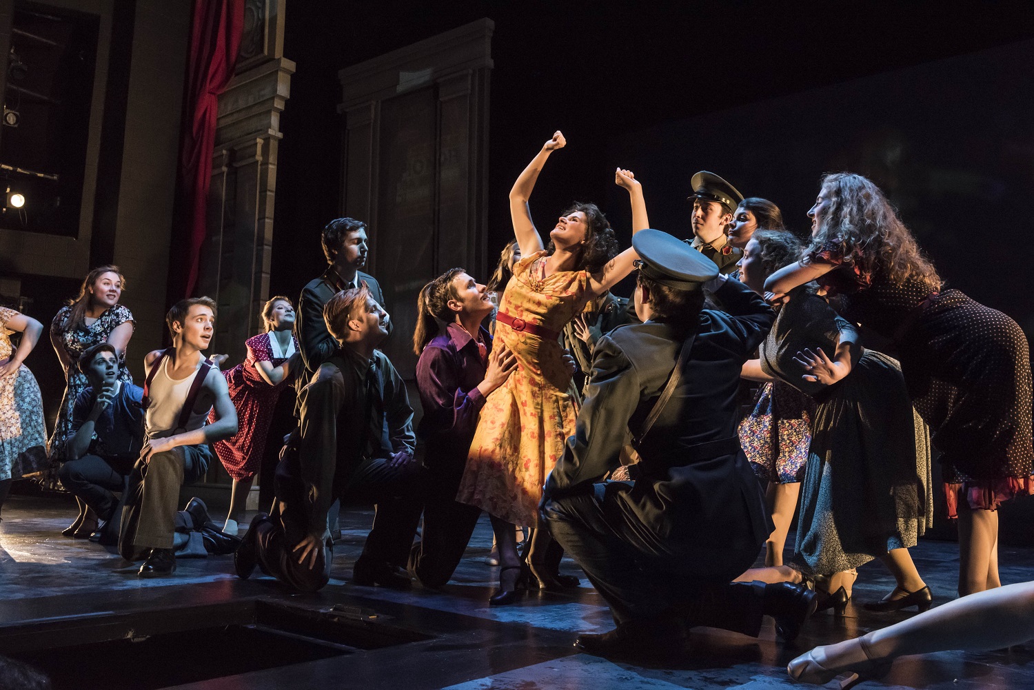 a large group students on stage performing Evita, there is dramatic lighting and one woman in the center with a spotlight on her face